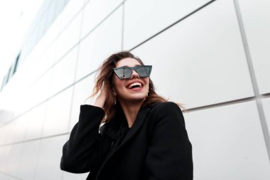 Gri bir binanın yakınındaki bir şehirde poz lüks bir siyah ceket bir gülümseme ile koyu şık güneş gözlüğü güzel neşeli genç hipster kadın. Çekici Avrupa neşeli kız açık havada gülüyor.