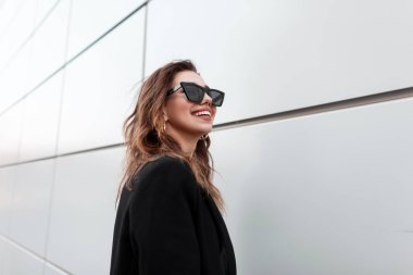 Şık bir siyah ceket güneş gözlüğü bir gülümseme ile Pozitif neşeli genç hipster kadın sıcak bir bahar gününde modern gri duvarın yanında duruyor. Neşeli mutlu kız modeli açık havada rahatlatır.