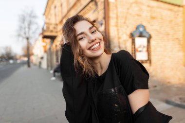 Şık bir tişörtle, şık bir ceketle, şehrin güneşli bir gününde klasik tuğla binaların yanında poz veren neşeli, güzel bir kadın. Yürüyüşe çıkmış neşeli bir kız. Bahar.