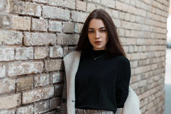 Pretty ung brunett kvinna med en naturlig make-up i en fashionabel pläd jacka med vit päls i en svart T-shirt vilar stående nära en tegel Vintage hus i staden. Snygg vacker flicka. — Stockfoto