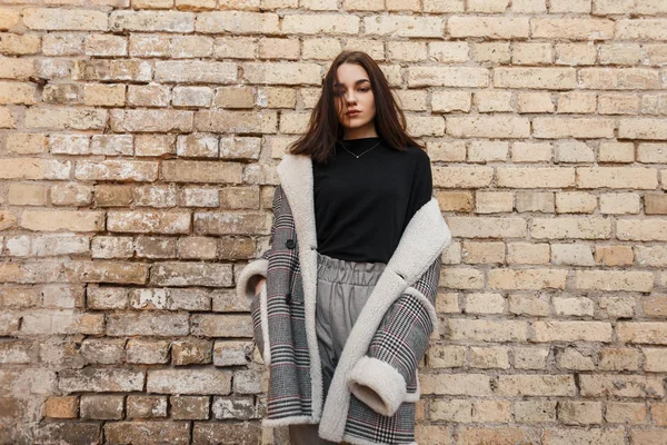 Pretty sexy young woman in a trendy black T-shirt in a luxurious checkered coat in gray stylish pants poses near a brick vintage wall outdoors. Modern fashionable brunette girl. Retro style. — Stock Photo, Image