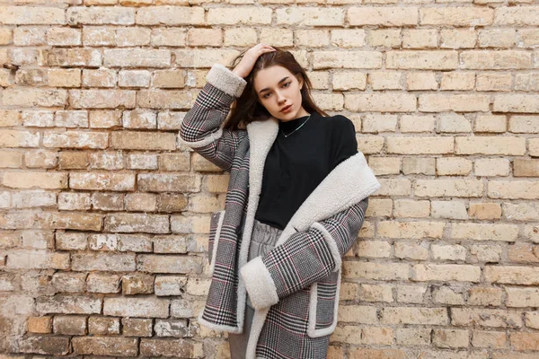 Cute pretty young woman in elegant checkered coat with white fur in a black stylish t-shirt in vintage gray pants posing near a brick wall. Fashionable european girl on a walk. Youth style. — Stock Photo, Image