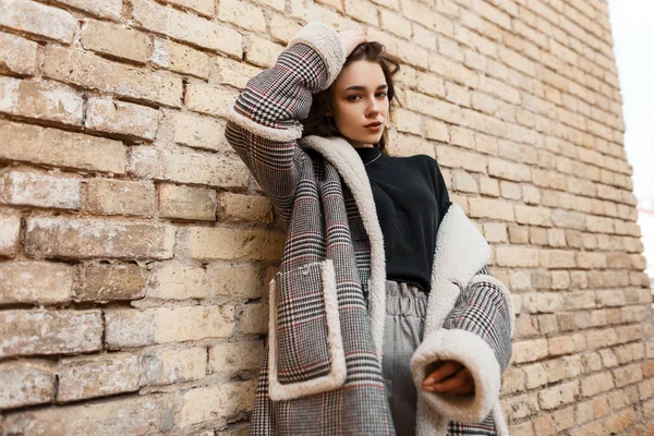 Attractive pretty young woman in trendy checkered coat with white fur in a black stylish t-shirt in vintage gray pants posing near a brick wall. Fashionable european girl on a walk. Modern youth style — Stock Photo, Image