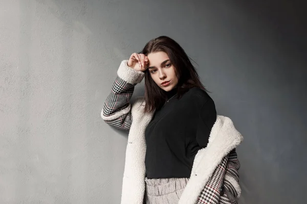 Young beautiful woman in a fashionable black T-shirt in a luxurious plaid jacket in elegant vintage pants is standing on the street near the concrete wall. Attractive girl brunette. Youth style. — Stock Photo, Image