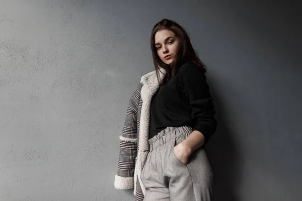 Fashionable modern young brunette woman in a black shirt in a gray stylish checkered jacket in gray vintage pants is standing in a room near a vintage concrete wall. Beautiful girl relaxes indoors. — Stock Photo, Image