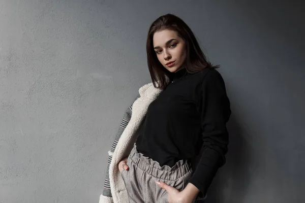 Mujer morena joven sexy europea en camisa negra en pantalones grises en chaqueta a cuadros gris de moda posando de pie cerca de una pared de hormigón vintage en el estudio. Elegante chica con estilo. Moda femenina . — Foto de Stock