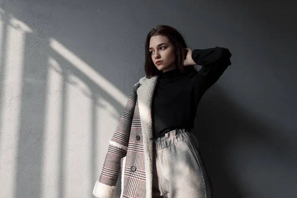 Modern stylish young woman in a fashionable shirt in a stylish checkered gray jacket in vintage pants stands on a sunny spring day indoors. Sensual beautiful brunette girl resting in the sunny room. — Stock Photo, Image