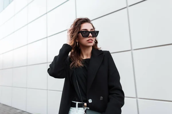 Mujer joven hipster bastante atractiva con un abrigo negro de moda en gafas de sol oscuras con estilo en jeans blancos en una elegante camiseta posa cerca de un edificio moderno al aire libre. Hermosa chica urbana. Estilo de calle . — Foto de Stock
