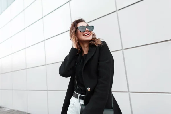 Vrolijke jonge vrouw hipster in zonnebrillen in een trendy jas in een T-shirt met een stijlvolle lederen handtas met een schattige glimlach in Vintage jeans staat in de buurt van een modern grijs gebouw in de stad. Gelukkig meisje — Stockfoto
