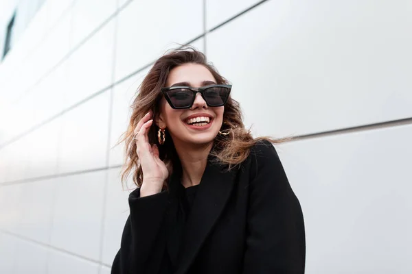 Портрет веселого Європейського молодого хіпстера жінки в стильних модних темних сонцезахисних окулярів в модному пальто в позитивній посмішки біля сірої стіни в місті. Міських щаслива дівчина насолоджується прогулянкою в Весняний день. — стокове фото