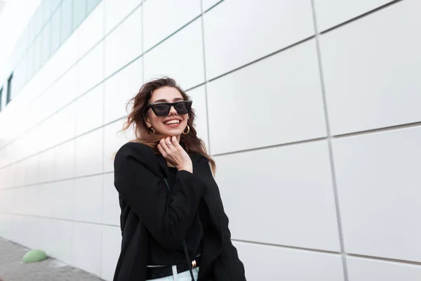 Drôle belle jeune femme hipster dans un manteau élégant noir en jean blanc vintage dans des lunettes de soleil sombres à la mode va et rit près d'un bâtiment gris dans le jour du printemps. positive joyeuse fille bénéficie d'une promenade . — Photo