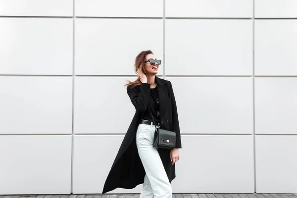 Jeune femme européenne joyeuse et hipster dans un long manteau noir vintage en jean blanc avec un élégant sac à main en cuir dans des lunettes de soleil sombres pose près d'un mur blanc moderne. Joyeux fille mignonne sur une promenade . — Photo