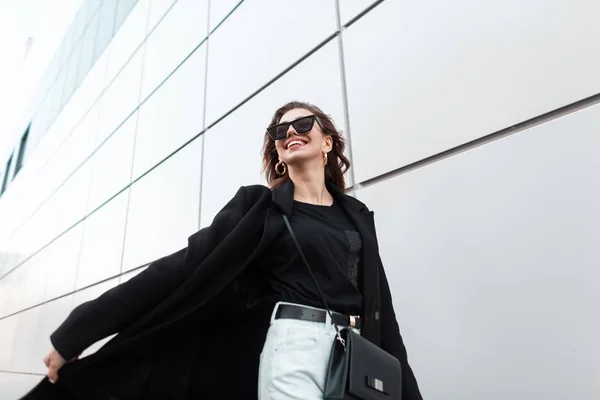 Mulher hipster jovem feliz em casaco preto longo na moda em jeans brancos em óculos de sol elegantes com uma bolsa de couro viaja ao redor da cidade perto da parede moderna. Menina positiva viajando pela rua . — Fotografia de Stock
