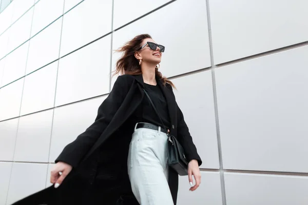 Modern glad ung hipster kvinna med ett positivt leende i solglasögon i svarta trendiga kläder reser runt i staden på en vårdag. Joyful Urban Girl njuter av helgen. Gatumode. — Stockfoto