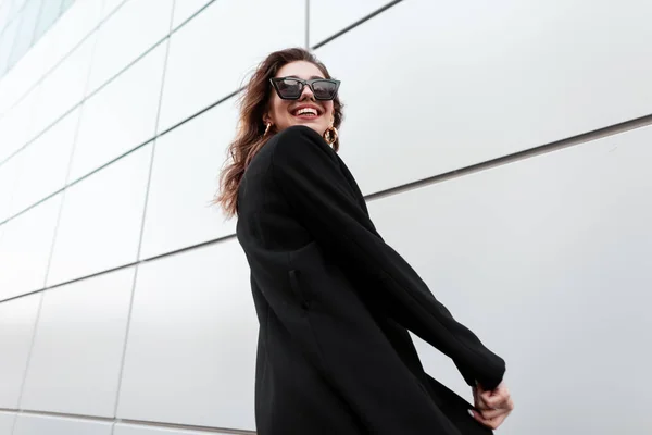 Jolie jolie belle femme hipster dans des lunettes de soleil élégantes dans un long manteau noir vintage avec un sourire positif pose et rit tout en se tenant près d'un bâtiment blanc moderne à l'extérieur. modèle fille heureuse . — Photo