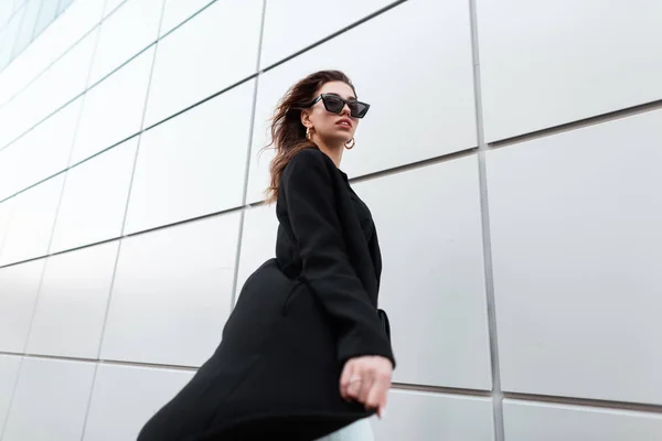 Jolie jeune femme hipster chic dans un long manteau noir chic dans des lunettes de style sombre posant sur une chaude journée de printemps près du mur moderne. Fille attrayant urbain marchant dans la rue. Style rétro . — Photo