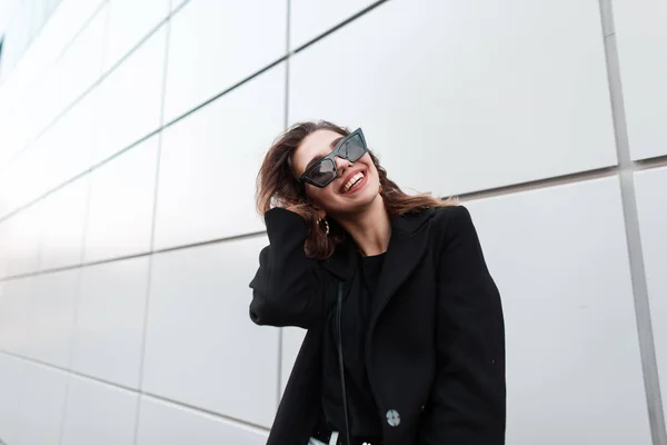 Jeune et drôle belle femme hipster en lunettes de soleil dans un manteau noir vintage dans un T-shirt avec un sourire positif pose et rit tout en se tenant près d'un bâtiment gris moderne dans la ville. Joyeuse fille . — Photo