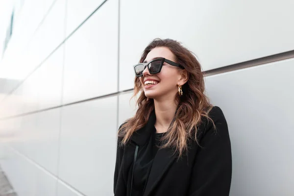 Jolie jeune femme hipster en noir élégant manteau élégant en lunettes de soleil est souriant debout près d'un mur moderne gris dans la ville. Joyeuse fille modèle aime marcher dans le jour du printemps. Mode de rue . — Photo