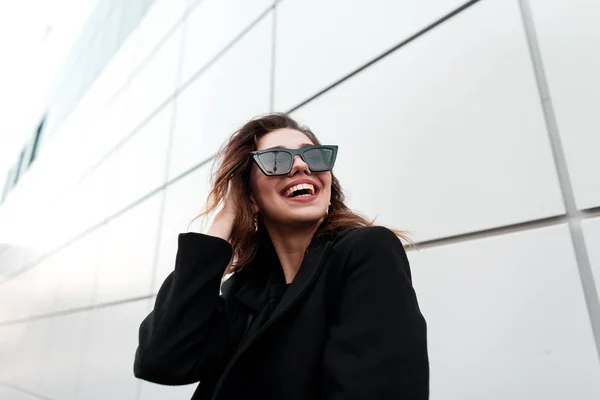 Belle jeune femme hipster joyeuse dans des lunettes de soleil sombres et élégantes avec un sourire dans un manteau noir luxueux posant dans une ville près d'un bâtiment gris. attrayant européen gai fille rire à l'extérieur . — Photo