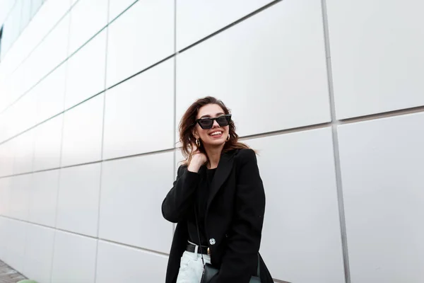 Jolie jeune femme hipster dans des lunettes de soleil sombres élégantes dans un élégant manteau noir avec un sac en cuir en jeans posant et souriant près du mur moderne de la ville. Modèle de fille gaie urbaine . — Photo