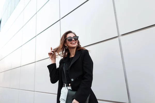 Drôle de jeune femme hipster dans un élégant manteau noir dans un T-shirt avec un sac à main en cuir à la mode avec un sourire positif en jean vintage est debout près d'un bâtiment gris moderne dans la ville. Joyeuse fille . — Photo