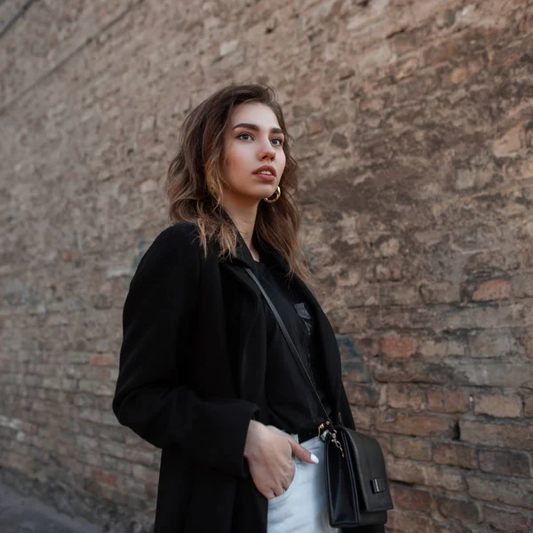 Mujer joven hermosa moderna con un abrigo elegante en una camiseta de moda en jeans blancos con una bolsa de cuero posando cerca de un antiguo edificio de ladrillo al aire libre. Hermosa chica disfruta del día de primavera. Estilo juvenil . — Foto de Stock