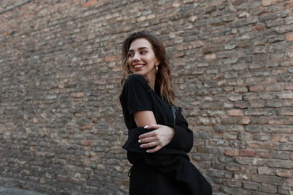 Hermosa joven feliz con una sonrisa atractiva en una elegante camiseta negra en un abrigo de moda posando cerca de una pared de ladrillo vintage al aire libre el día de primavera. Positivo alegre chica modelo disfruta del fin de semana . — Foto de Stock