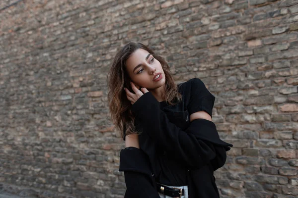 Bastante sexy modelo de moda de mujer joven en un elegante abrigo negro en una camiseta de moda está posando cerca de un edificio de ladrillo vintage marrón. Elegante chica atractiva al aire libre en la ciudad. Estilo moderno . — Foto de Stock