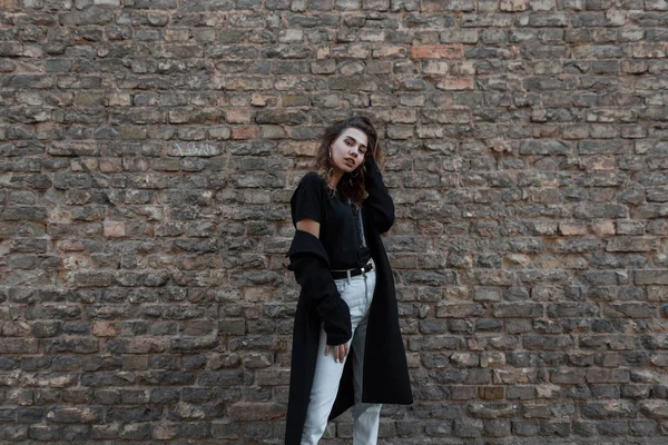 Europeisk snygg ung kvinna i en Vintage svart lång kappa i vita jeans i en fashionabel t-shirt poserar stående nära tegel brun vägg utomhus. Ganska attraktiv tjej för en promenad. Dammode — Stockfoto
