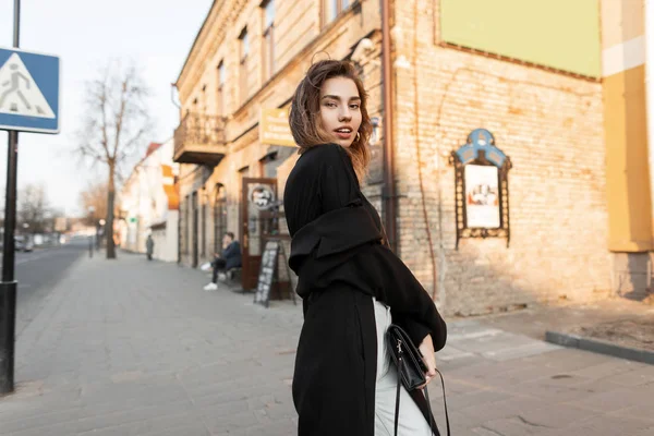 Elegante mujer joven moderna con un largo abrigo vintage con un bolso de cuero negro en un vaquero blanco de moda en una camiseta caminando por la calle en un día soleado. Hermosa chica modelo relaja al atardecer . — Foto de Stock