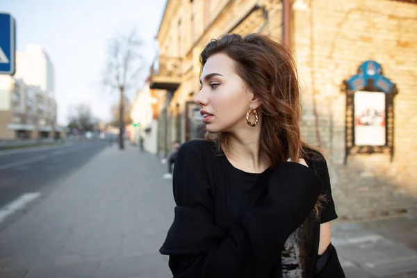 Jolie jolie jeune femme dans un T-shirt noir à la mode dans un manteau élégant se promène dans la ville près des bâtiments en brique vintage et regarde la route. Belle fille voyage au coucher du soleil. Printemps . — Photo