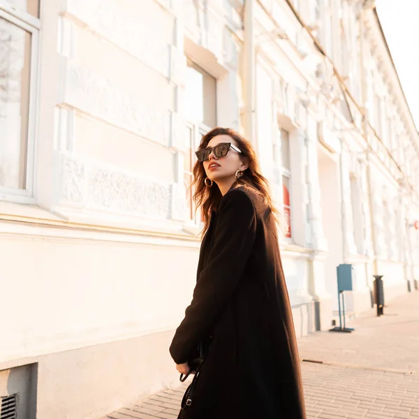 Moderna moderno jovem morena hipster mulher em preto elegante casaco longo em óculos de sol elegantes posando perto de um edifício branco em um dia ensolarado. Modelo de moda menina europeia viaja ao redor da cidade . — Fotografia de Stock