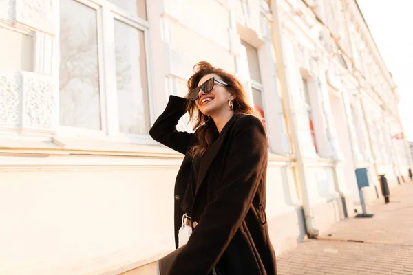 Jeune femme heureuse hipster dans des lunettes de soleil noires avec un beau sourire dans des vêtements de printemps à la mode se promène le long de la rue dans la ville et profite d'une journée chaude et ensoleillée. Joyeuse fille modèle en plein air . — Photo
