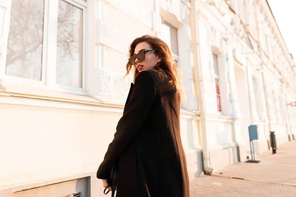 Mulher hipster muito jovem europeu em casaco preto na moda em óculos de sol elegantes com uma bolsa de couro está de pé em uma rua na cidade em um dia de primavera ensolarado brilhante. Menina atraente ao ar livre . — Fotografia de Stock