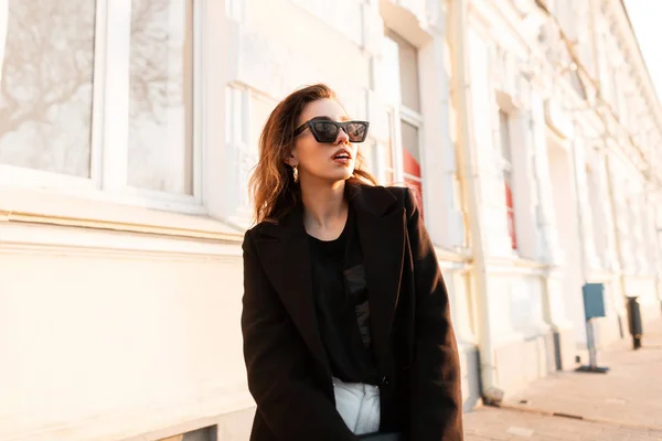 Mooie elegante hipster jonge vrouw in trendy zonnebrillen in een zwart modieus t-shirt in een stijlvolle jas in jeans staat op een zonnige dag in de buurt van een historische straat in de stad. Aantrekkelijk modern meisje. — Stockfoto