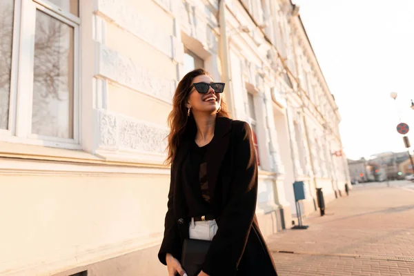 Aantrekkelijke jonge hipster vrouw met een positieve glimlach in een modieuze jas in zonnebrillen in een T-shirt in jeans met een handtas staat in de stad en geniet van het felle zonlicht. Vrij gelukkig meisje. — Stockfoto