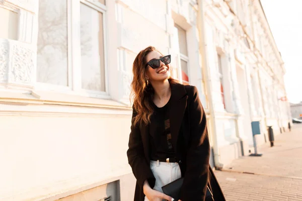 Grappige vrolijke hipster jonge vrouw in stijlvolle zonnebril met trendy jas in witte jeans met een handtas wandelen op straat in de buurt van vintage gebouwen. Gelukkig aantrekkelijk meisje reist bij zonsondergang. — Stockfoto