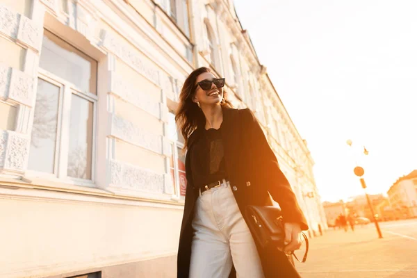 Modieuze positieve jonge hipster vrouw in een chique lange vacht in donkere zonnebrillen in een t-shirt in witte jeans met een leren tas op de achtergrond van een oranje zonsondergang in de stad. Gelukkig meisje buitenshuis. — Stockfoto