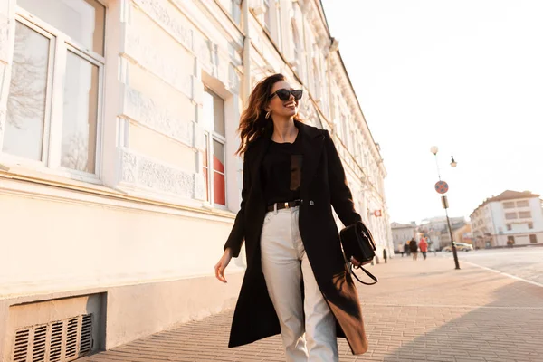 Muito feliz jovem hipster mulher em jeans da moda com uma bolsa de couro em óculos de sol elegantes em um casaco longo preto caminha pelas ruas da cidade perto de um edifício vintage em um dia ensolarado. Menina alegre . — Fotografia de Stock