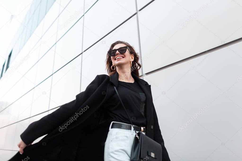 Happy young hipster woman in fashionable long black coat in white jeans in stylish sunglasses with a leather bag travels around the city near the modern wall. Positive girl traveling down the street.