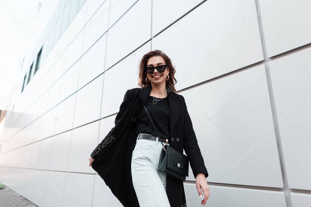 Positive young stylish hipster woman in sunglasses in an elegant coat in a T-shirt in vintage jeans with a leather handbag is walking and smiling near a modern building in the city.Cheerful girl walks
