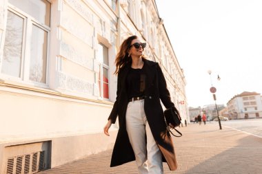 Siyah uzun ceket şık güneş gözlüğü bir deri çanta ile trendy kot Pretty mutlu genç hipster kadın güneşli bir günde bir vintage binanın yakınında şehir sokaklarında yürür. Neşeli kız.