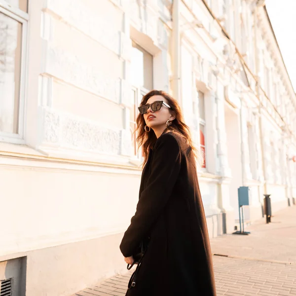 Mode moderne jeune femme brune hipster en noir élégant manteau long dans des lunettes de soleil élégantes posant près d'un bâtiment blanc par une journée ensoleillée. modèle de mode fille européenne voyage autour de la ville . — Photo