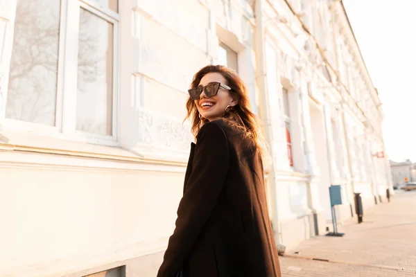 Joven mujer hipster alegre con una sonrisa positiva en gafas de sol negras en un elegante abrigo posando cerca de un edificio vintage blanco en la ciudad en un día soleado brillante. Chica alegre modelo disfruta de la puesta del sol . — Foto de Stock