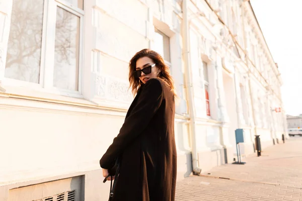 Jovem mulher hipster muito elegante em casaco preto vintage em óculos escuros da moda está posando ao ar livre em um dia ensolarado brilhante perto de um edifício moderno. Menina atraente caminha pelas ruas da cidade . — Fotografia de Stock