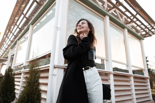 Mulher bonita feliz em uma camiseta preta na moda em um casaco longo luxuoso em jeans vintage com um sorriso bonito está posando na rua perto de um café de verão. Urban modelo menina alegre goza de uma caminhada . — Fotografia de Stock