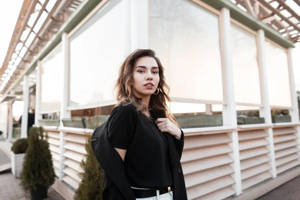 Urban όμορφη νεαρή γυναίκα με ένα μαύρο παλτό σε ένα μοντέρνο t-shirt σε λευκό τζιν με μια πόζα που ποζάρει κοντά σε ένα vintage ξύλινο κτίριο σε εξωτερικούς χώρους. Ευρωπαϊκό μοντέλο κορίτσι απολαμβάνει την ημέρα της άνοιξης. Στυλ νεολαίας. — Φωτογραφία Αρχείου
