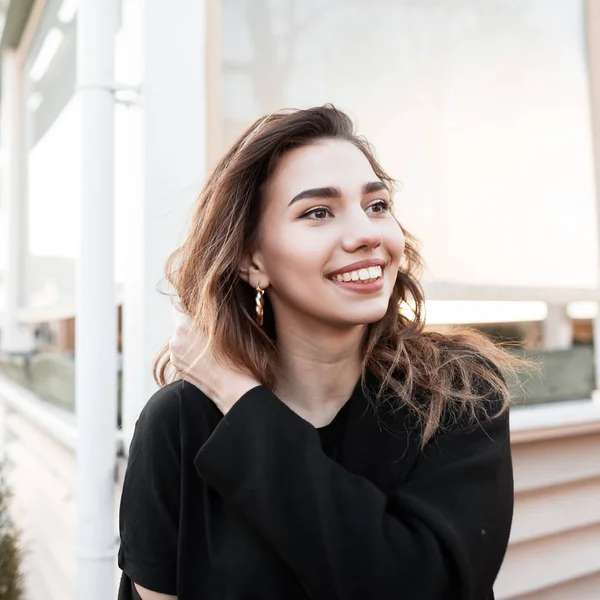 Portrét veselé mladé ženy s atraktivním úsměvem v módním černém kabátě nedaleko dřevěné letní kavárny. Pozitivní radostná dívka v šťastném jarním dnu. Moderní styl mládeže. — Stock fotografie