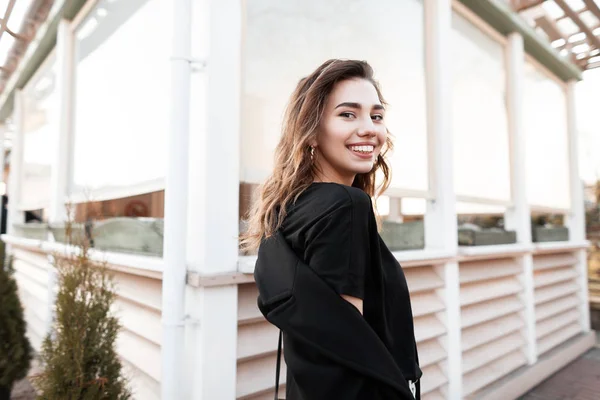 Mulher bonita bonita com um sorriso atraente bonito em uma camiseta preta em um casaco preto na moda posando em pé na rua perto de um edifício de madeira. Menina feliz positivo goza de fim de semana de primavera — Fotografia de Stock