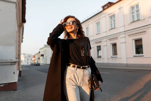 Mulher hipster jovem alegre na moda em um casaco longo em óculos escuros em uma camiseta em calça branca com uma bolsa de couro no fundo de um pôr-do-sol laranja na cidade. Menina feliz bonito ao ar livre . — Fotografia de Stock